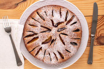 Sweet homemade plum cake with white ceramic dish, knife and fork on wooden table, macro, top view.
