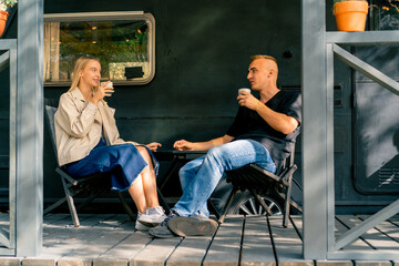 A young couple a man and a girl are drinking tea on the terrace near their motorhome in camper