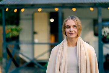 Portrait of a young beautiful smiling girl with a blanket on her shoulders against background of a mobile home in glamping - obrazy, fototapety, plakaty