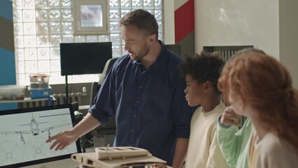 Medium shot of Caucasian male engineering teacher showing airplane drawing scheme on desktop computer to multiracial primary school kids in classroom