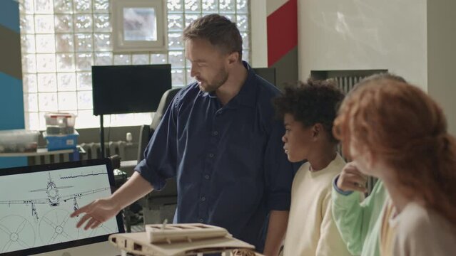 Medium shot of Caucasian male engineering teacher showing airplane drawing scheme on desktop computer to multiracial primary school kids in classroom