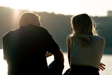 Silhouette of couple in love sitting on the sandy bank of the river admiring the setting sun and...