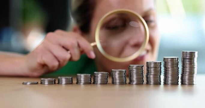 A woman with a magnifying glass looks at the coins in a row on the table, close-up. Investment growth