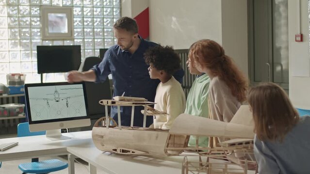 Medium shot of diverse group of classmates listening to Caucasian male teacher showing airplane construction plan on computer desktop during engineering lesson in classroom