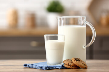 Glass and jug of fresh milk with sweet cookies on wooden table in kitchen