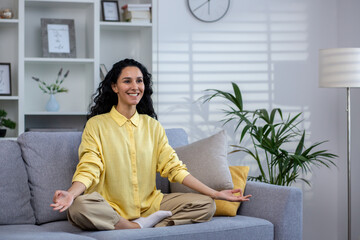 Young beautiful woman meditating at home sitting on sofa in living room in lotus pose, latin...