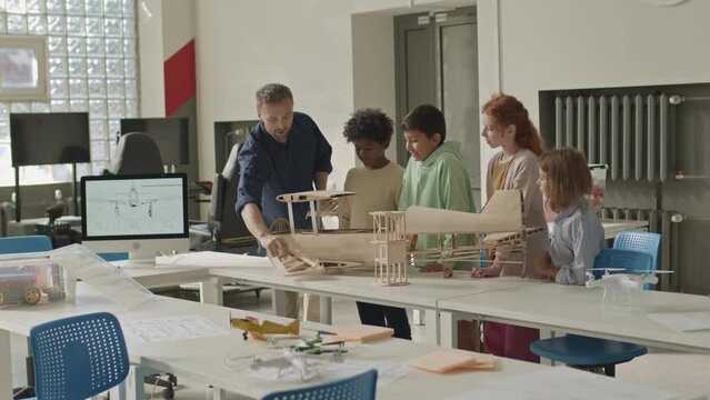 Medium long shot of Caucasian male teacher showing wood handmade airplane construction model on desk in classroom to group of multiracial primary school children attending engineering club together