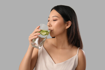 Beautiful Asian woman drinking cucumber water on grey background, closeup