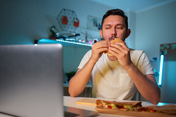 Guy with a beard biting a hamburger while sitting in a dining room at home