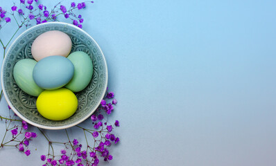 Multicolored pastel green, blue eggs lie in round gray plate on blue background. Easter composition top view. Easter eggs flatly with copy space. Spring season flowers. Easter postcard place for text.