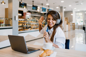 Beautiful and happy young woman sitting and eating delicious rolls in bakery or fast food. She also...