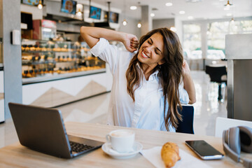 Beautiful and happy young woman sitting and eating delicious rolls in bakery or fast food. She also...