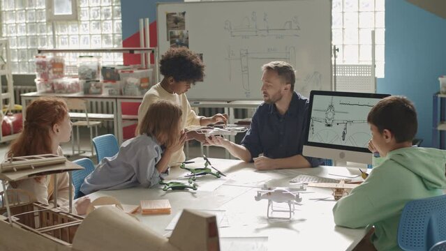 Medium shot of group of multiracial preteen schoolkids making aircraft models and showing them to Caucasian male teacher during engineering lesson in spacious classroom at daytime