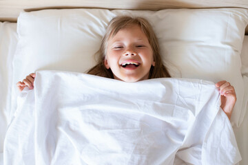 A charming happy little girl with long hair is lies on a white bed under a white blanket and laughs...