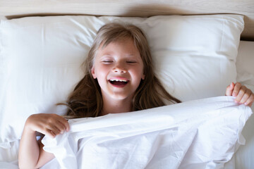 Obraz na płótnie Canvas A charming happy little girl with long hair is lies on a white bed under a white blanket and laughs at the morning. It's perfect for projects related to family, childhood, or relaxation.Top view photo
