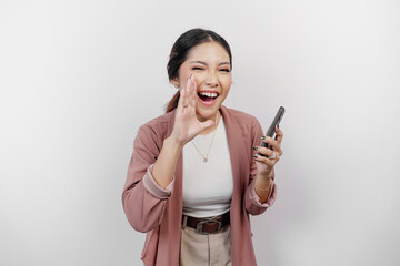 A young beautiful Asian woman employee wearing cardigan and holding her phone is shouting and screaming loud with a hand on her mouth, isolated by white background.