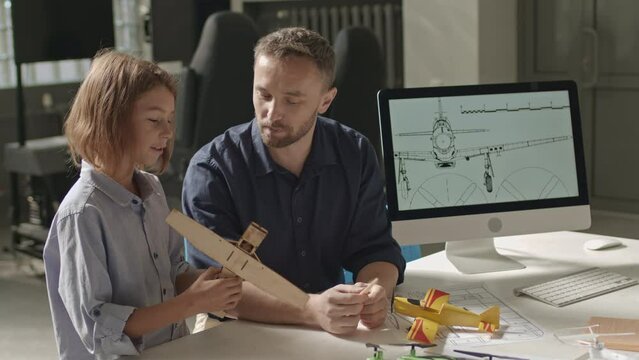 Medium shot of Caucasian elementary age school boy showing wooden handmade plane model to young male teacher in engineering club