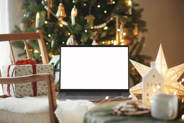 Naklejka na ściany i meble Laptop with empty screen and stylish christmas gifts on modern chair in festive decorated room with tree, golden lights and star. Christmas shopping online and sales. Laptop mock up, copy space