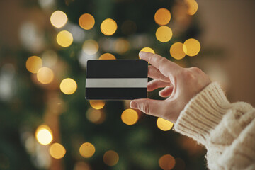 Hand in cozy sweater holding credit card close up on background of christmas tree lights in festive...
