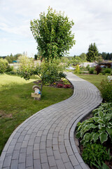 Curved Pathway from gray bricks in the garden. Decorative Landscaping Design. Winding brick paving path to house. Flowers, shrubs and garden sculpture near the path.