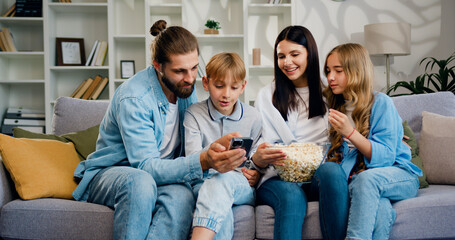 Parents father mother and cute little kids children watch funny social media video on mobile phone. Young family using modern wireless tech watching vlog, enjoy free time on internet.