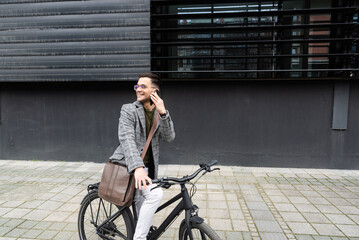 Happy employee environment lover calling colleagues before staff meeting. Cheerful businessman with glasses on reading text messages on a smart phone and pushing a bicycle.
