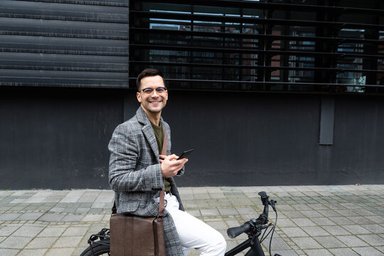 Happy employee environment lover calling colleagues before staff meeting. Cheerful businessman with glasses on reading text messages on a smart phone and pushing a bicycle.