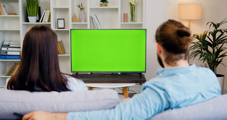 Happy couple watching tv and switching channels looking at green screen in modern living room. Woman and man watching chroma key television. - obrazy, fototapety, plakaty
