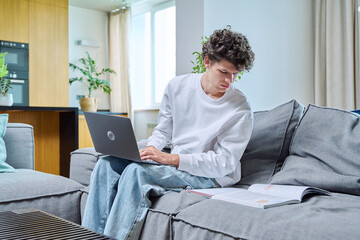 Handsome guy college student studying at home typing on laptop, sitting on sofa