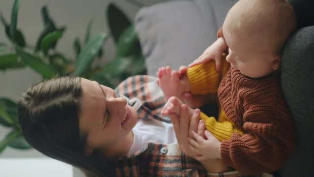 Vertical video of happy young loving mom gently kiss little feet sitting on cozy couch at home, tiny toes of newborn baby child. Tender moments with kid, mother and offspring connection, care concept