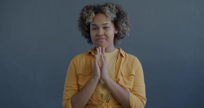 Slow motion portrait of evil young African American woman planning thinking rubbing hands looking at camera on grey color background
