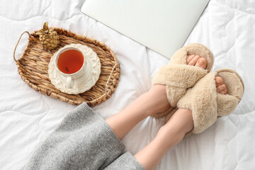 Young woman in soft slippers resting on bed with modern laptop and cup of tea - Powered by Adobe