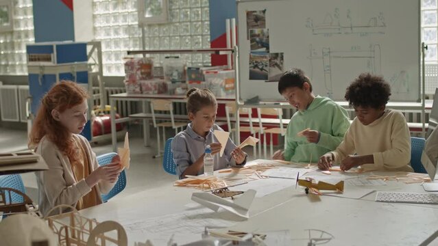 Medium shot of ethnically diverse school kids making wood plane models at work desk in spacious technical and engineering classroom