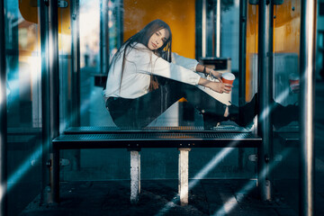 young woman with a glass of coffee sitting at a bus stop