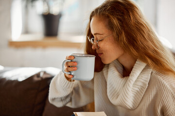 Young woman with pleasant appearance and freckles drinks hot coffee, tea in cozy home environment. Nice quiet weekend. concept of lifestyle, winter holiday season, autumn weekend, relax atmosphere.