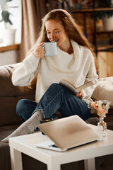 Obraz premium Young girl in freckles, with stylish glasses and book in her hand. She drinks fragrant warming tea or coffee while sitting at home on sofa. concept of lifestyle, winter holiday season, relax.