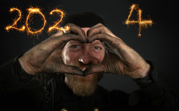 Schornsteinfeger in Arbeitskleidung blickt durch seine zu einem Herz geformten Hände, Jahreszahlen 2024  mit Sternspritzer geschrieben, dunkler Hintergrund.