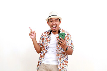 Excited asian adult man tourist holding a cell phone while pointing sideways. Concept of travel. Isolated on white background