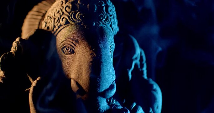 Ganesha idol made of sandstone on a black background at night. There is warm light and blue light. There is smoke from the incense that people burn to worship.
