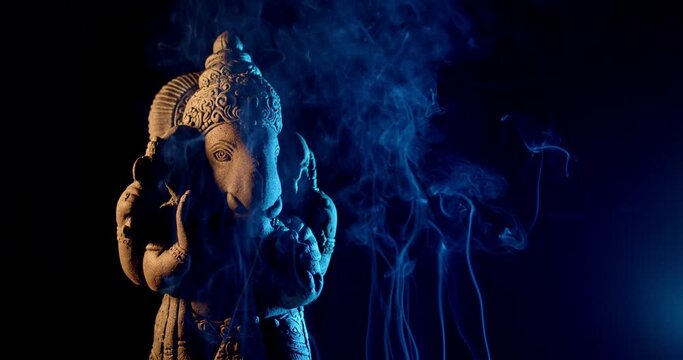 Ganesha idol made of sandstone on a black background at night. There is warm light and blue light. There is smoke from the incense that people burn to worship.