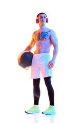 Shirtless young man with fit, muscular, relief body standing in headphones with fitness ball over white studio background in neon light. Concept of sport, active and healthy lifestyle, body, fitness