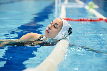 Focus on young restful female swimmer in cap standing in water against finish line and looking...