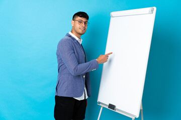 Young Colombian man isolated on blue background giving a presentation on white board and writing in...