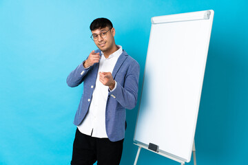 Young Colombian man isolated on blue background giving a presentation on white board and points...
