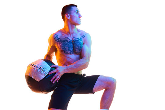 Muscular, sportive young man with strong, relief shirtless body doing lunges with fitness ball against white studio background in neon light. Concept of sport, active and healthy lifestyle, fitness