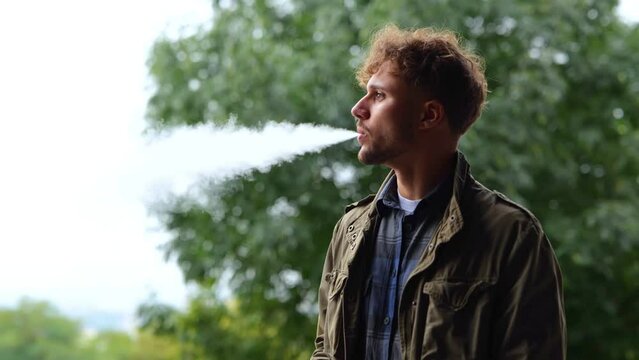 Side view of Caucasian young handsome man smokes a vape outside. Casual thoughtful male smoking electronic cigarette while standing outdoors in park looking away. Real time concept.