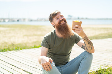 Redhead man with beard holding fried chips at outdoors looking up while smiling