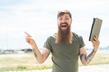 Redhead man with beard holding a tablet at outdoors surprised and pointing finger to the side