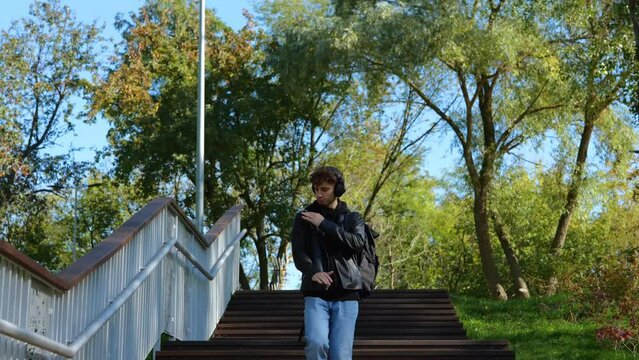 Stylish young man going down stairs and listening the music with headphones in the park while dancing. Lifestyle. Concept of autumn walks. Real time concept.