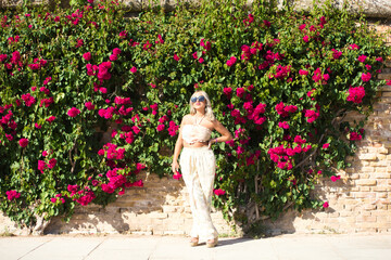 Pretty young blonde woman wearing sunglasses and dressed in embroidered trousers and crocheted top, enjoys sunny day. In the background and on the wall a large pink bougainvillea plant.